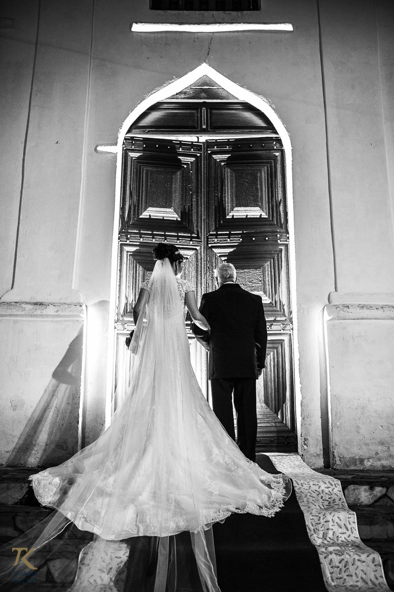 Casamento de Sabrina e Alisson. Noiva espera a hora de entrar na igreja junto a seu pai. Portas fechadas, foto em preto e branco.