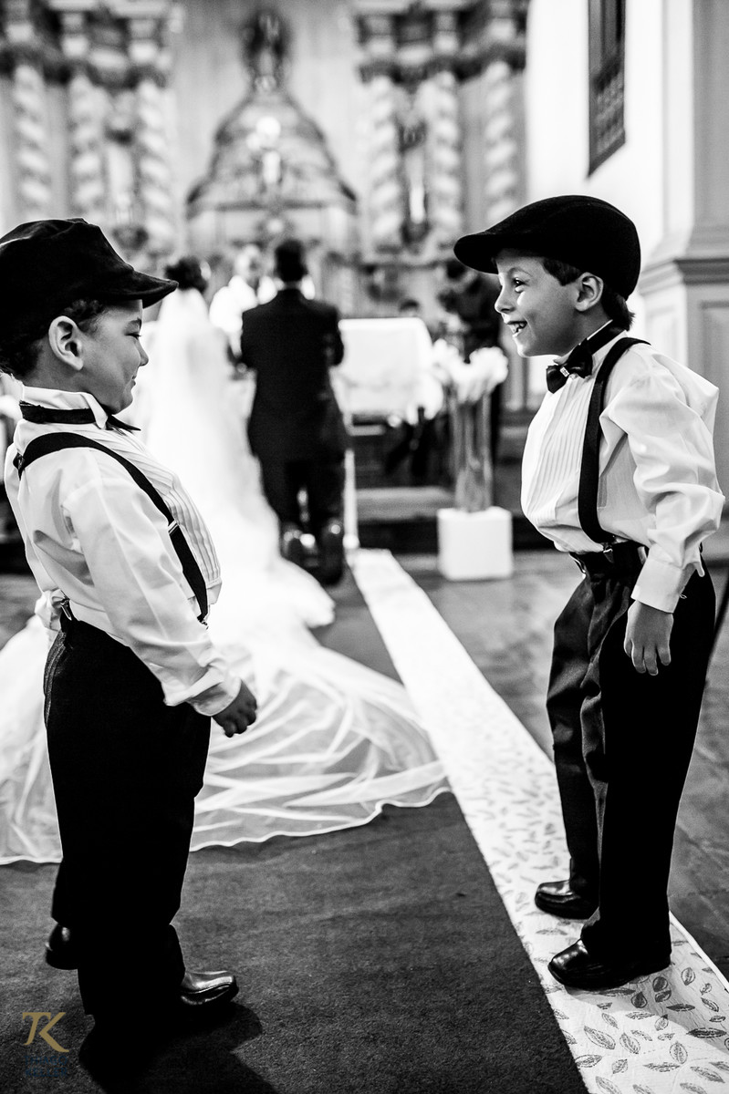 Casamento de Sabrina e Alisson. Pajens brincam no corredor da IIgreja e ao fundo os noivos estão ajoelhados.
