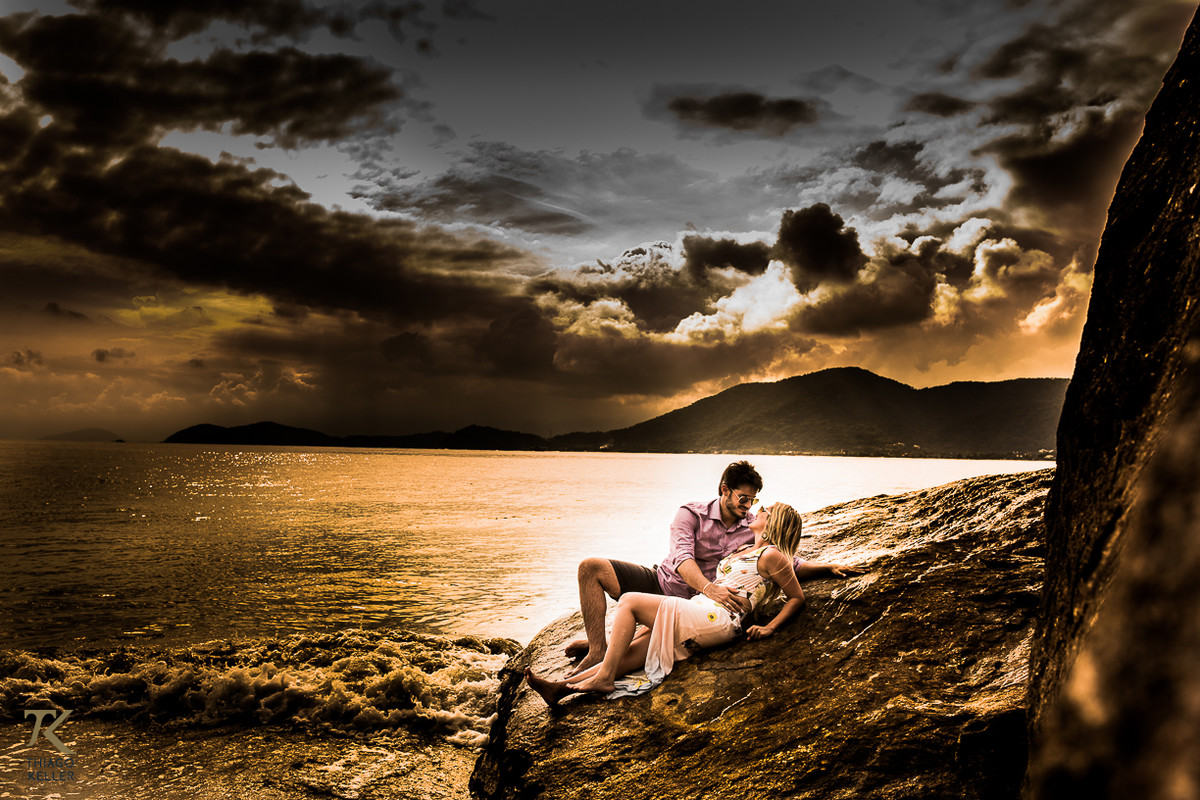 Casal deitado em uma rocha, se olhando e abraçados com o mar ao fundo.