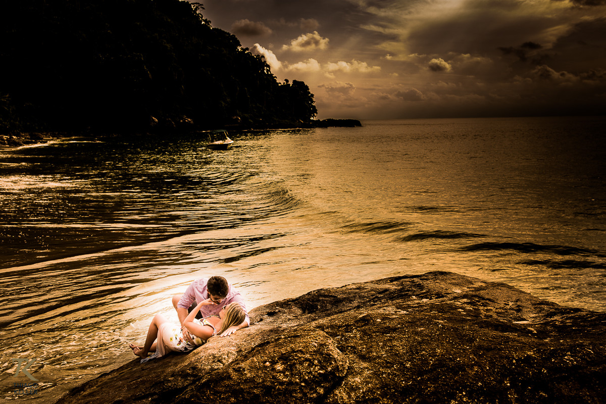 Casal deitado em uma rocha, se olhando e abraçados com o mar ao fundo.