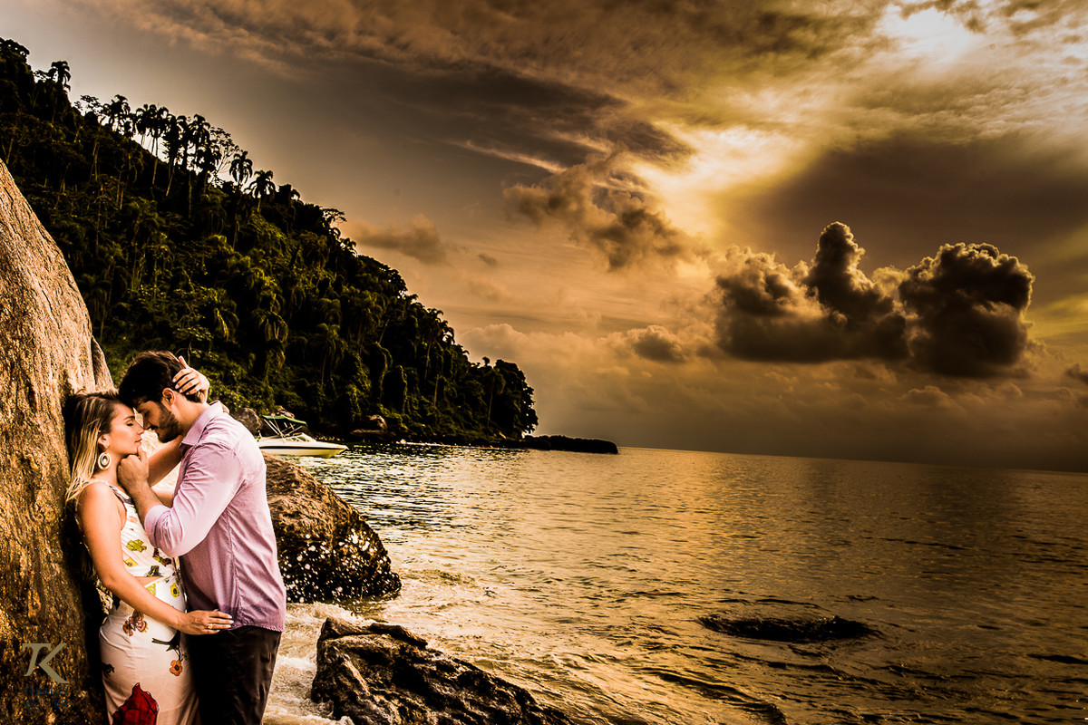 Casal abraçado, conectado um ao outro, ao fundo o mar e o sol, escondido por trás das nuvens traz um tom dourado à foto.