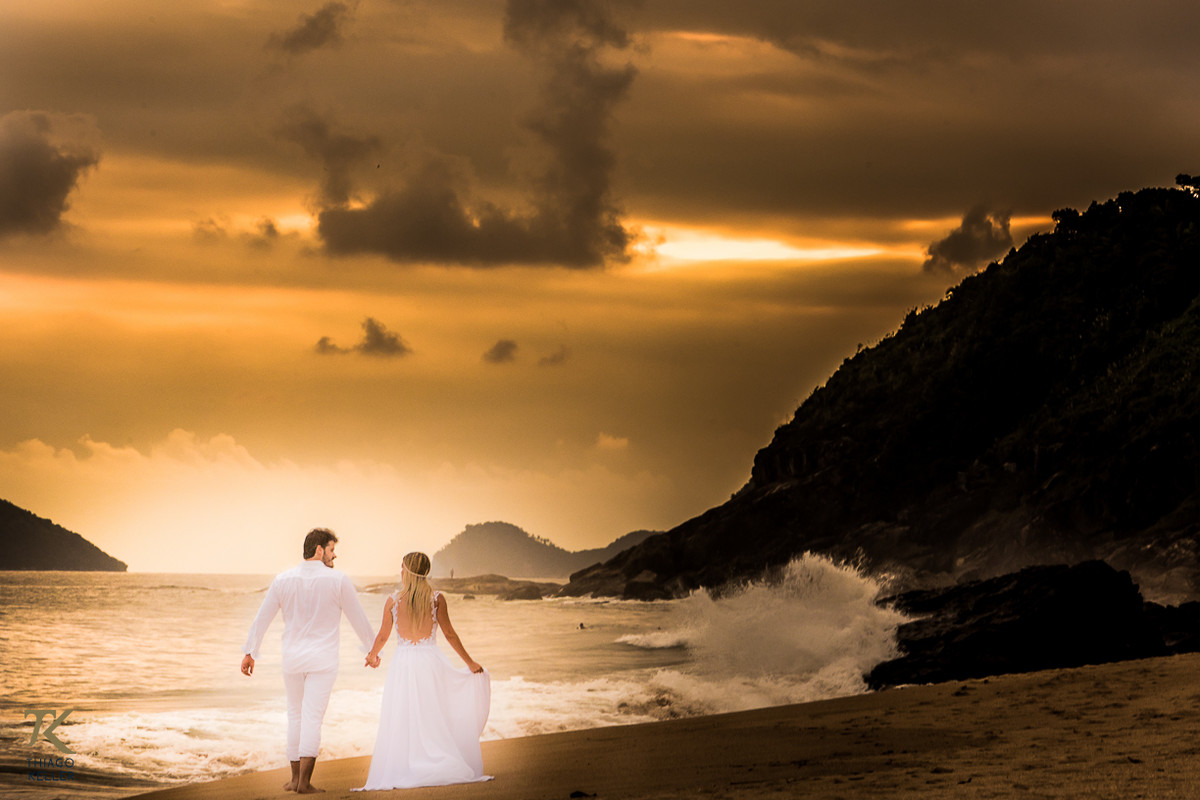 Casal caminha vestido de branco em direção do por do sol na praia.