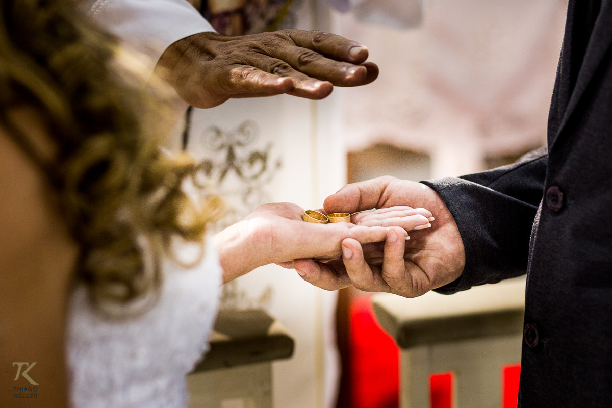 Fotografia de casamento de Lívia e Paulo Henrique na cidade de Paracatu, Minas Gerais. Padre abençoa as alianças do casal