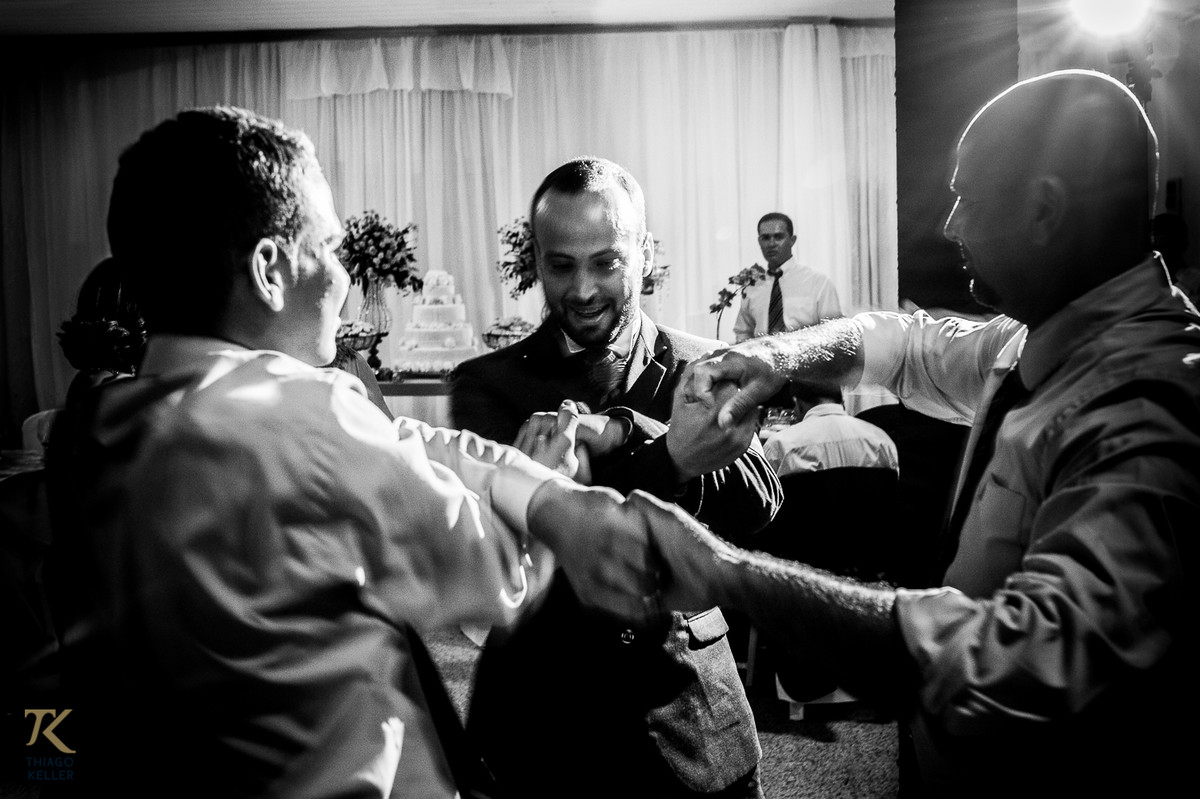 Fotografia de casamento de Lívia e Paulo Henrique na cidade de Paracatu, Minas Gerais. O noivo, seu pai e seu irmão, dançam de mãos dadas.