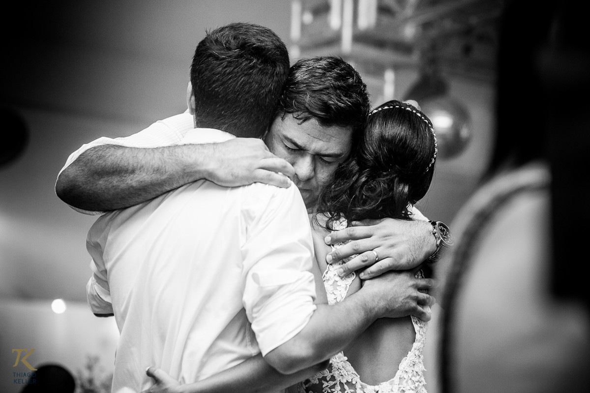 Fotografia de Casamento de Maura e Thiago realizado na cidade de Paracatu, Minas Gerais. Casal é abraçado por amigo durante a festa. Foto em preto e branco.