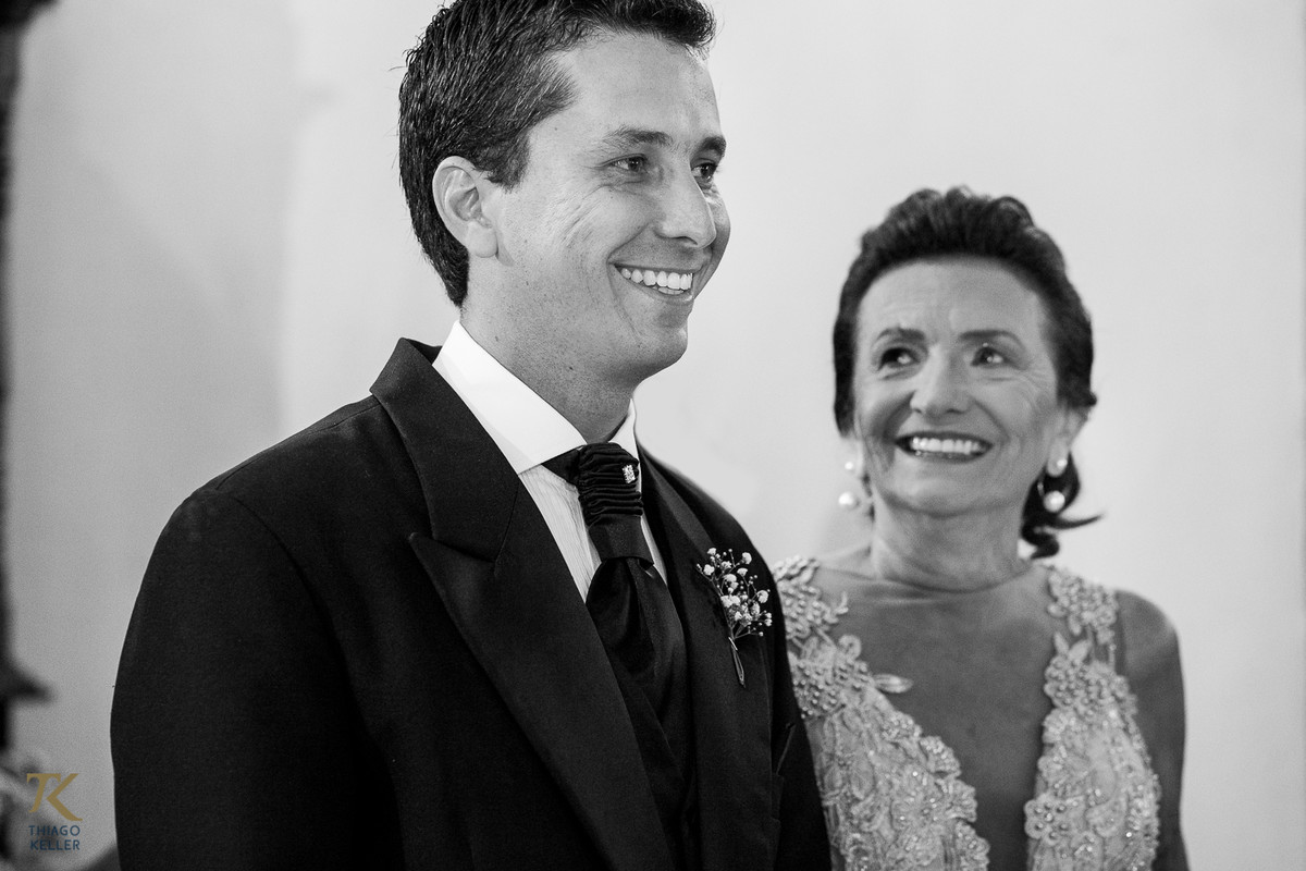 Fotografia de Casamento de Maura e Thiago realizado na cidade de Paracatu, Minas Gerais. Mãe do noivo sorri para noivo no altar.  Foto em preto e branco.