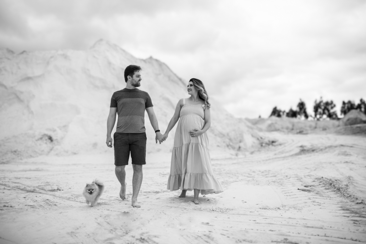 O casal caminhando nas dunas, lado a lado, com o cachorrinho acompanhando. A imagem evoca união, cumplicidade e a alegria de estarem juntos neste momento especial. Fotos feitas pelo fotógrafo de gestante Rafael Ohana em Brasília.