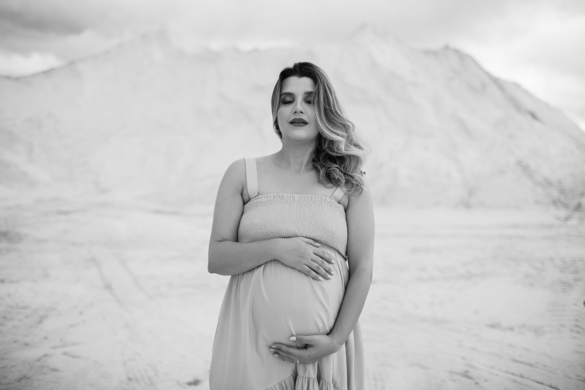 Close-up da gestante com as mãos sobre a barriga. A expressão tranquila e o cenário das dunas ao fundo realçam a força e delicadeza da maternidade. Fotos feitas pelo fotógrafo de gestante Rafael Ohana em Brasília.