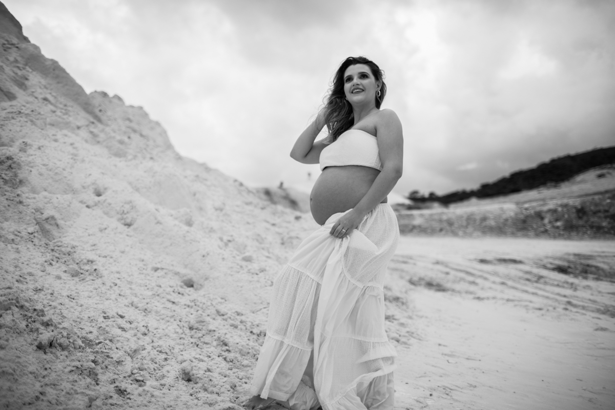 A gestante em uma pose contemplativa, de perfil, olhando para o horizonte. A vastidão das dunas cria um efeito de tranquilidade e eternidade na foto. Fotos feitas pelo fotógrafo de gestante Rafael Ohana em Brasília.