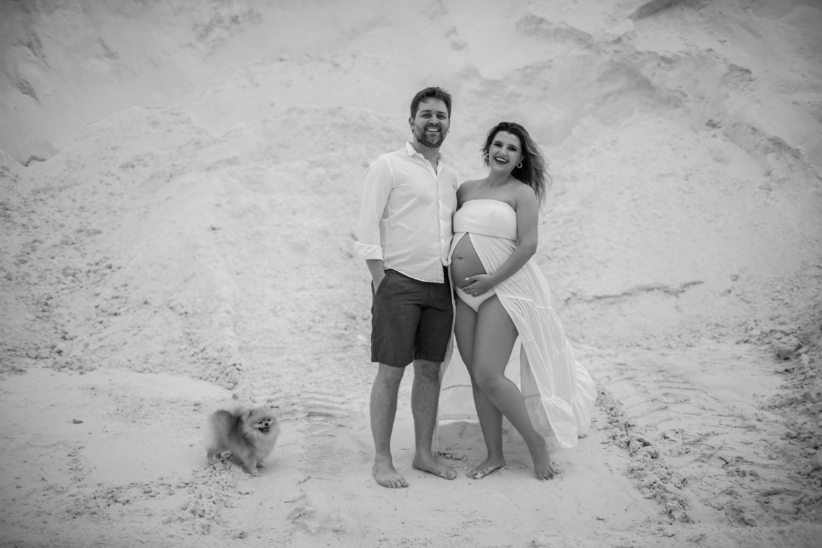 Foto do casal gestante de pé em meio às dunas, com o homem ao lado da gestante, criando um momento de proximidade e carinho. O cenário das dunas ao fundo destaca a beleza natural do local. Fotos feitas pelo fotógrafo de gestante Rafael Ohana em Brasília.