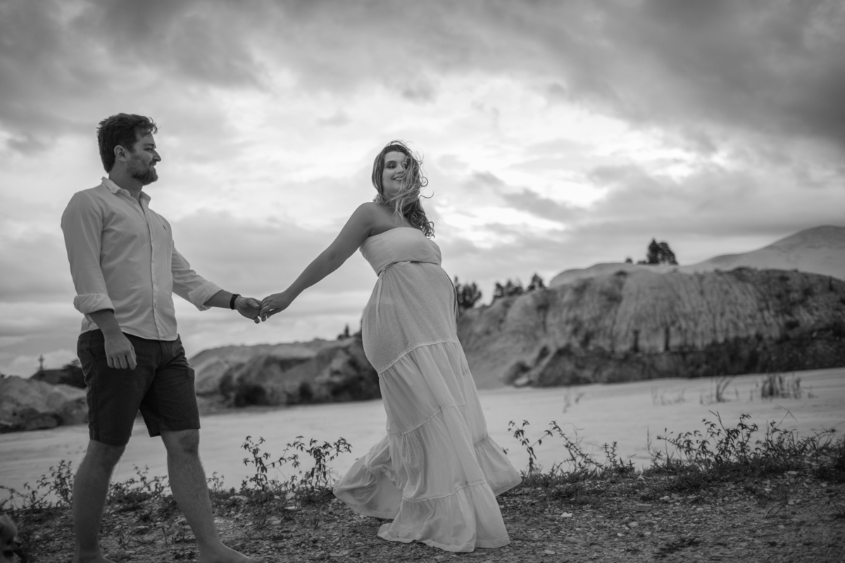 O casal andando de mãos dadas em um momento espontâneo e alegre, enquanto o vento sopra levemente o vestido da gestante, capturando a leveza e a conexão entre os dois. Fotos feitas pelo fotógrafo de gestante Rafael Ohana em Brasília.