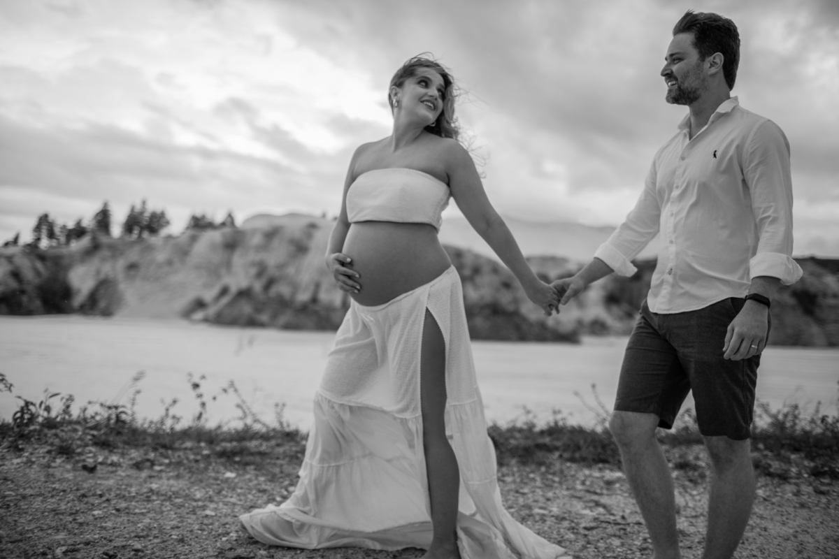 O casal andando de mãos dadas em um momento espontâneo e alegre, enquanto o vento sopra levemente o vestido da gestante, capturando a leveza e a conexão entre os dois. Fotos feitas pelo fotógrafo de gestante Rafael Ohana em Brasília.