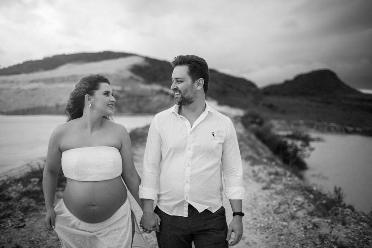 O casal se olhando de maneira carinhosa, com o homem de mãos nos bolsos e a gestante tocando a barriga. A conexão e a expectativa pelo bebê são evidentes, e a paisagem natural reforça a tranquilidade do momento. Fotos feitas pelo fotógrafo de gestante Raf