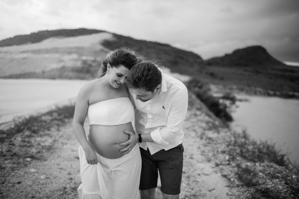 O casal se olhando de maneira carinhosa, com o homem de mãos nos bolsos e a gestante tocando a barriga. A conexão e a expectativa pelo bebê são evidentes, e a paisagem natural reforça a tranquilidade do momento. Fotos feitas pelo fotógrafo de gestante Raf