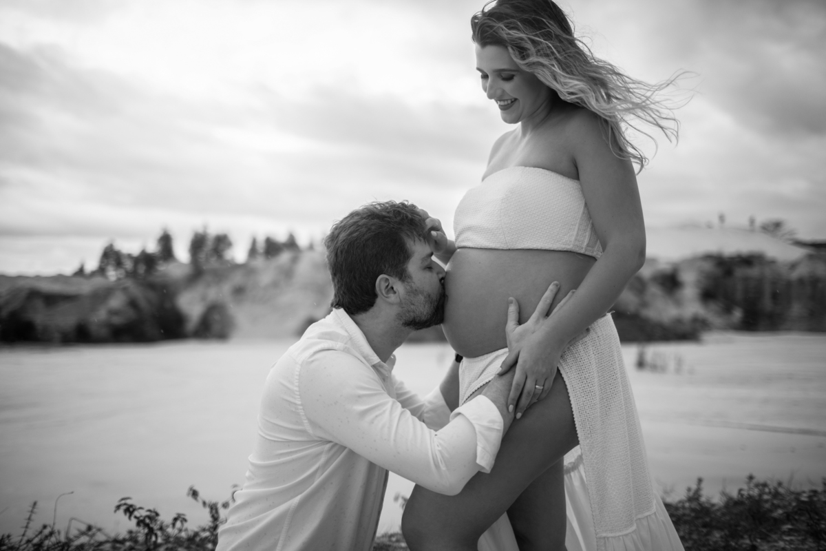 Cena romântica em que o homem beija a barriga da gestante, ajoelhado, enquanto ela sorri para a câmera. Um momento de carinho capturado em um ambiente natural ao ar livre. Fotos feitas pelo fotógrafo de gestante Rafael Ohana em Brasília.