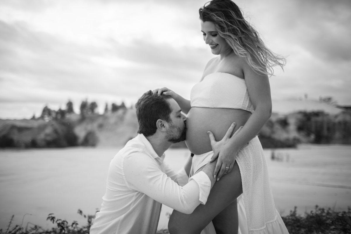 Cena romântica em que o homem beija a barriga da gestante, ajoelhado, enquanto ela sorri para a câmera. Um momento de carinho capturado em um ambiente natural ao ar livre. Fotos feitas pelo fotógrafo de gestante Rafael Ohana em Brasília.