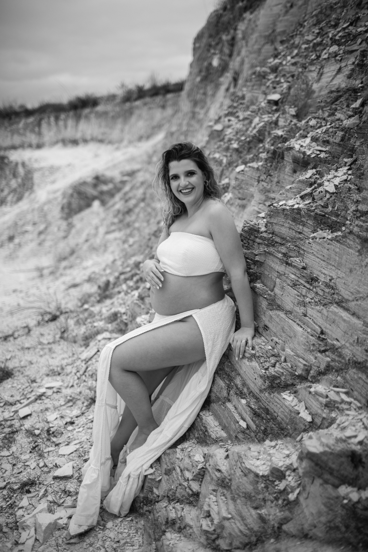 Close-up da gestante apoiada em uma rocha, com a mão sobre a barriga e o olhar pensativo. O contraste entre o corpo humano e a textura natural das pedras realça a beleza do momento. Fotos feitas pelo fotógrafo de gestante Rafael Ohana em Brasília.