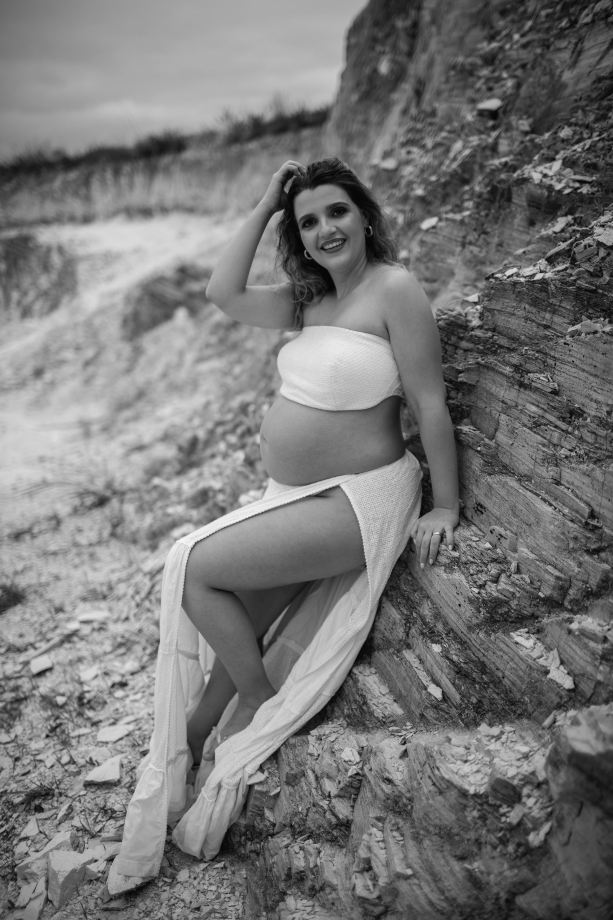 Close-up da gestante apoiada em uma rocha, com a mão sobre a barriga e o olhar pensativo. O contraste entre o corpo humano e a textura natural das pedras realça a beleza do momento. Fotos feitas pelo fotógrafo de gestante Rafael Ohana em Brasília.