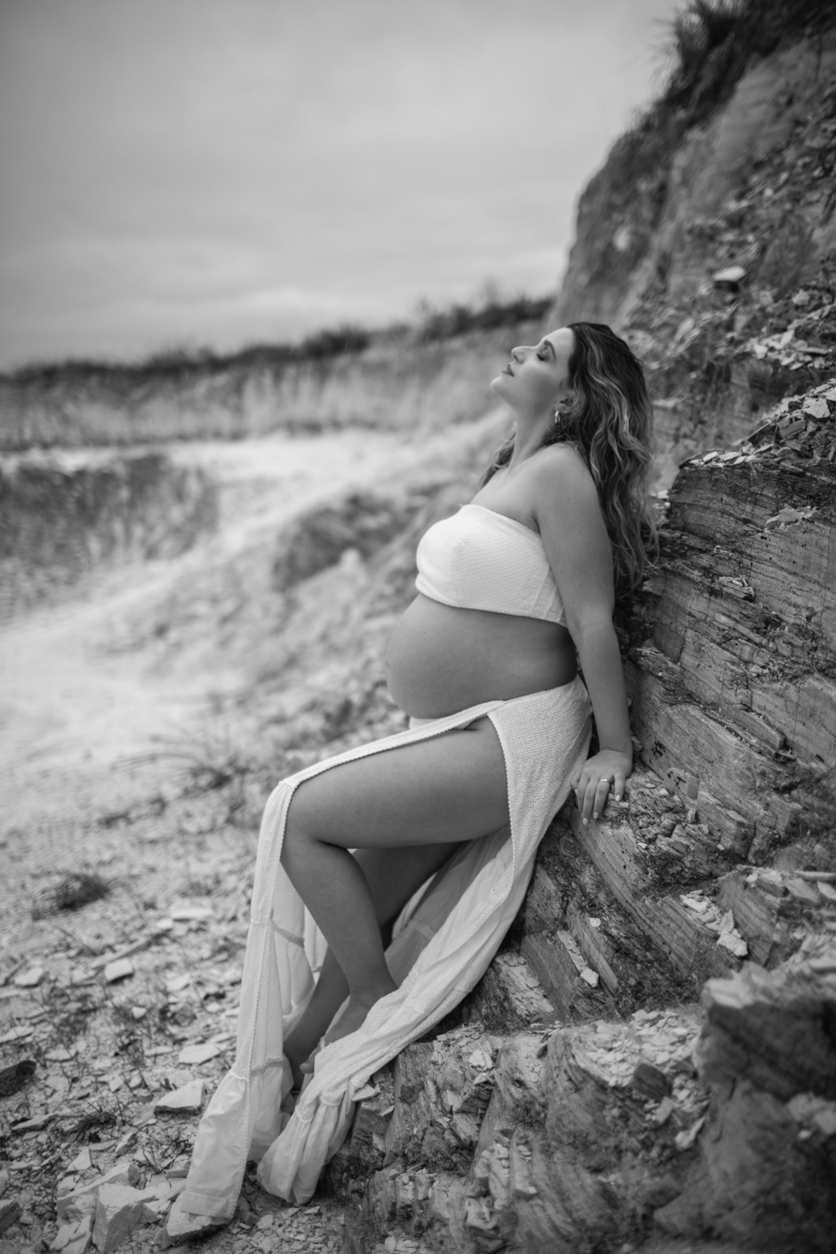 Close-up da gestante apoiada em uma rocha, com a mão sobre a barriga e o olhar pensativo. O contraste entre o corpo humano e a textura natural das pedras realça a beleza do momento. Fotos feitas pelo fotógrafo de gestante Rafael Ohana em Brasília.