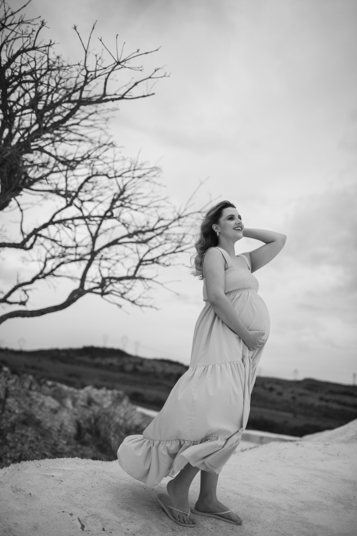 Outra perspectiva da mesma gestante com a árvore seca, capturada em um ângulo lateral, mostrando a delicadeza de sua silhueta. O ambiente continua a transmitir uma sensação de calmaria, mesmo com o céu carregado. Fotos feitas pelo fotógrafo de gestante Ra