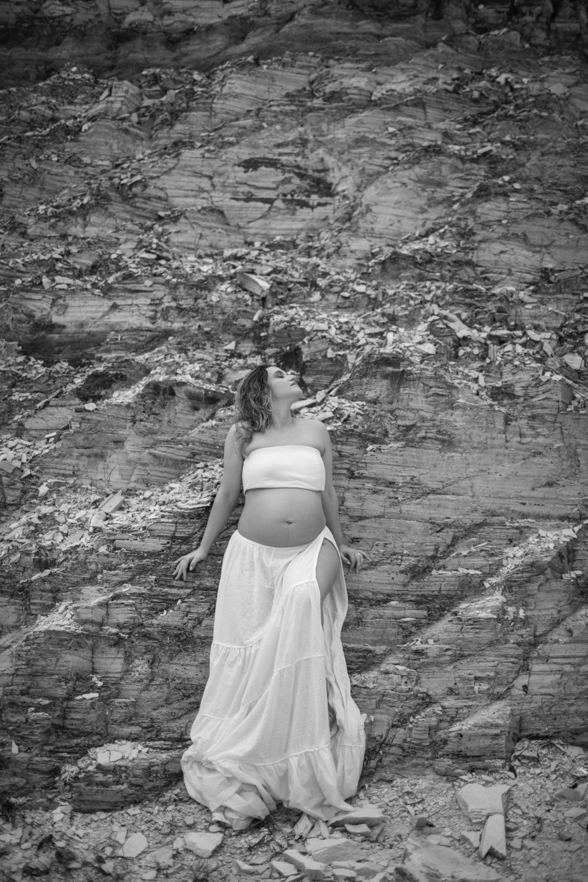 A gestante se encostando na rocha, com um leve sorriso no rosto, enquanto o vento sopra o seu vestido. A combinação de elementos naturais e o momento emocional criam uma imagem harmoniosa. Fotos feitas pelo fotógrafo de gestante Rafael Ohana em Brasília.
