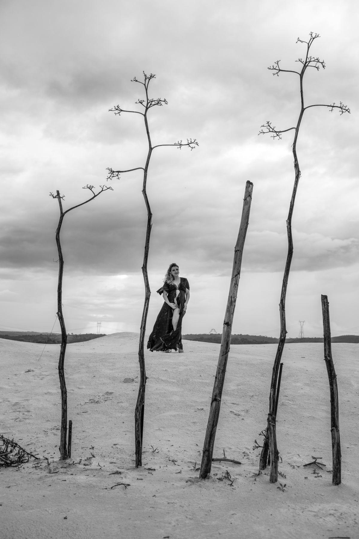 gestante em pé em frente a uma árvore seca com galhos nus, criando um contraste visual impactante entre a natureza árida e o florescer da vida. Fotos feitas pelo fotógrafo de gestante Rafael Ohana em Brasília.