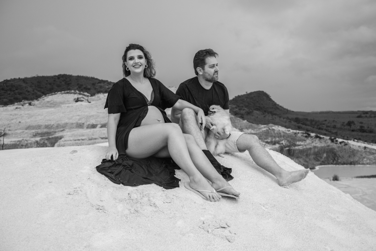 Cena familiar com o casal gestante e o cachorro sentados juntos na areia, olhando para o horizonte. O momento tranquilo reflete o carinho e a união da família. Fotos feitas pelo fotógrafo de gestante Rafael Ohana em Brasília.