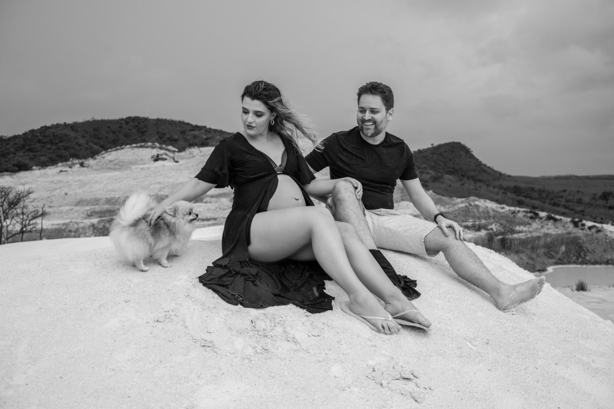 Cena familiar com o casal gestante e o cachorro sentados juntos na areia, olhando para o horizonte. O momento tranquilo reflete o carinho e a união da família. Fotos feitas pelo fotógrafo de gestante Rafael Ohana em Brasília.