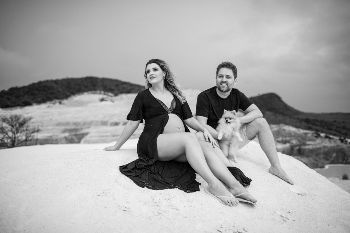 Cena familiar com o casal gestante e o cachorro sentados juntos na areia, olhando para o horizonte. O momento tranquilo reflete o carinho e a união da família. Fotos feitas pelo fotógrafo de gestante Rafael Ohana em Brasília.