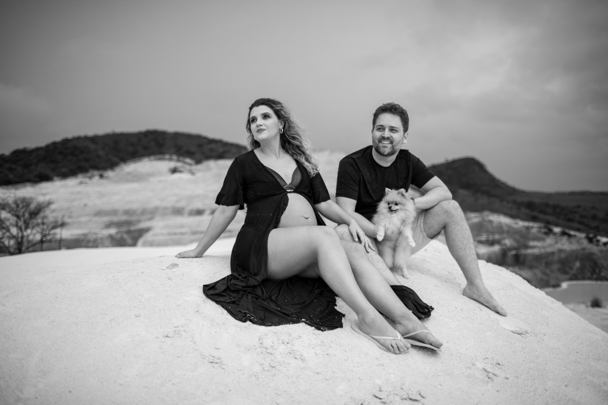 Cena familiar com o casal gestante e o cachorro sentados juntos na areia, olhando para o horizonte. O momento tranquilo reflete o carinho e a união da família. Fotos feitas pelo fotógrafo de gestante Rafael Ohana em Brasília.