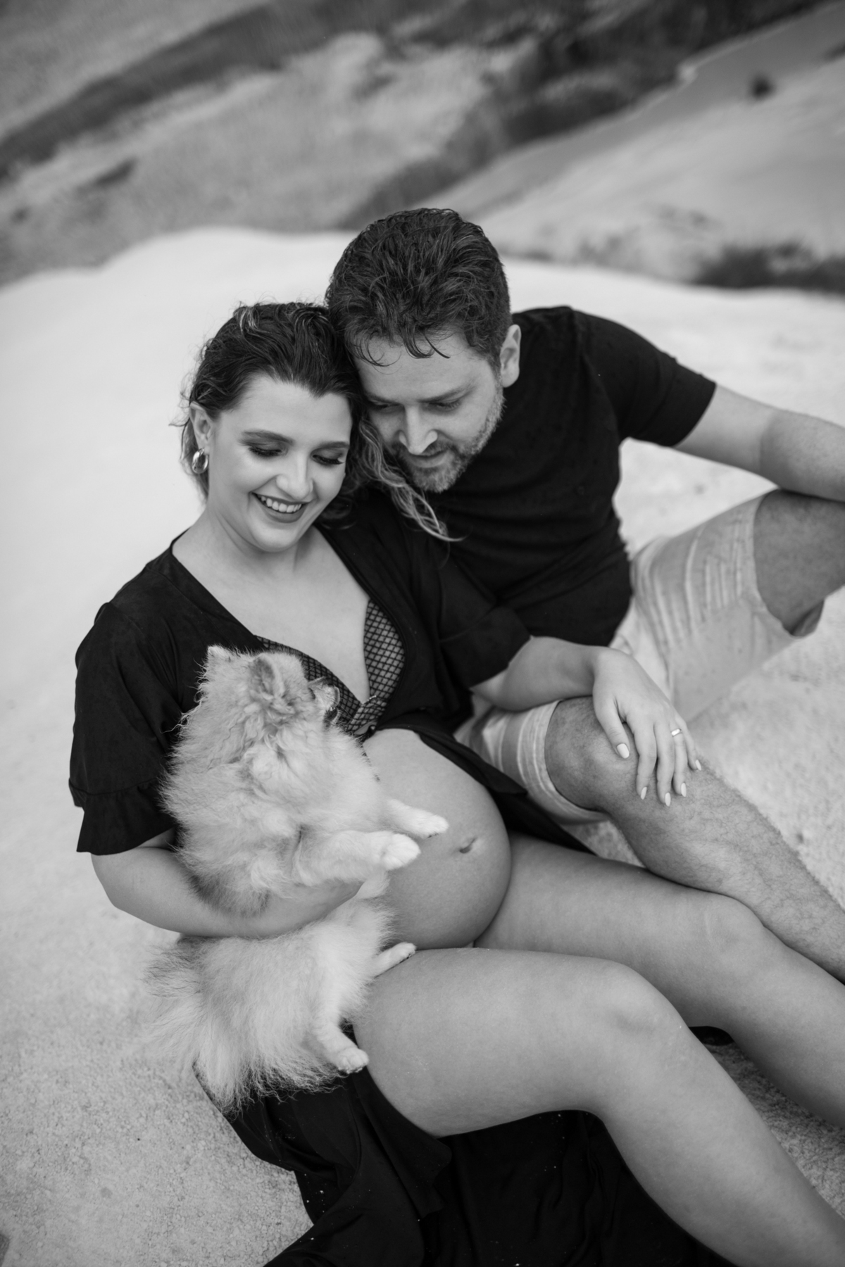 Cena familiar com o casal gestante e o cachorro sentados juntos na areia, olhando para o horizonte. O momento tranquilo reflete o carinho e a união da família. Fotos feitas pelo fotógrafo de gestante Rafael Ohana em Brasília.