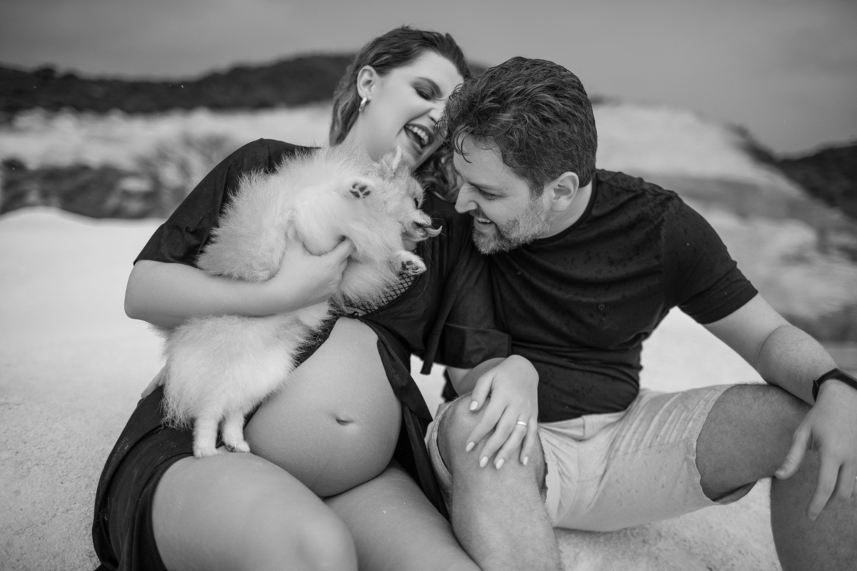 Cena familiar com o casal gestante e o cachorro sentados juntos na areia, olhando para o horizonte. O momento tranquilo reflete o carinho e a união da família. Fotos feitas pelo fotógrafo de gestante Rafael Ohana em Brasília.