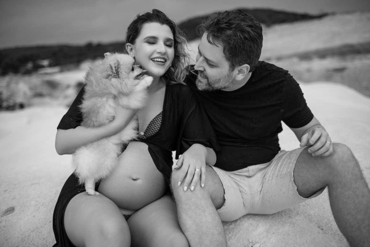Cena familiar com o casal gestante e o cachorro sentados juntos na areia, olhando para o horizonte. O momento tranquilo reflete o carinho e a união da família. Fotos feitas pelo fotógrafo de gestante Rafael Ohana em Brasília.