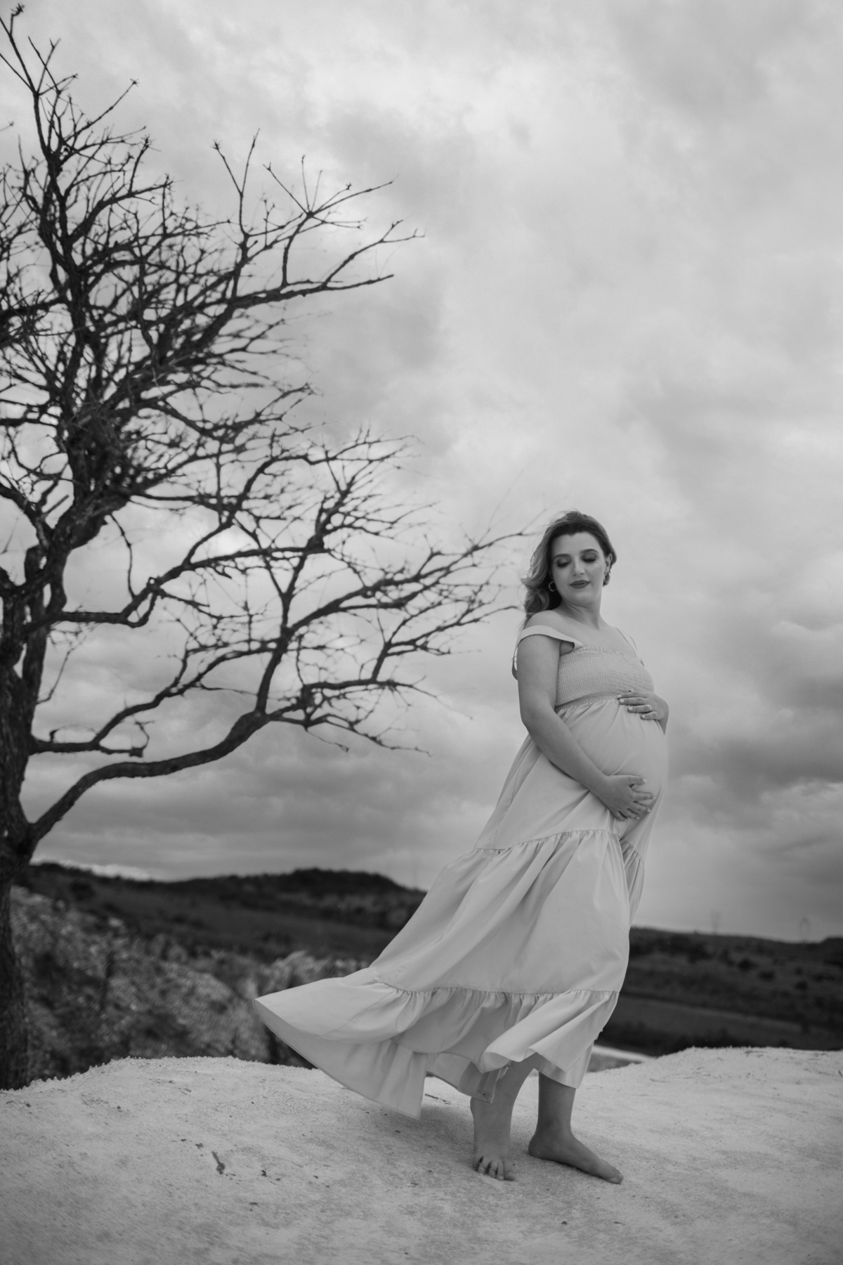 Um close-up da gestante, próxima à árvore, exibindo sua barriga com as mãos apoiadas. A expressão dela revela uma conexão profunda com o momento e o ambiente natural ao redor. Fotos feitas pelo fotógrafo de gestante Rafael Ohana em Brasília