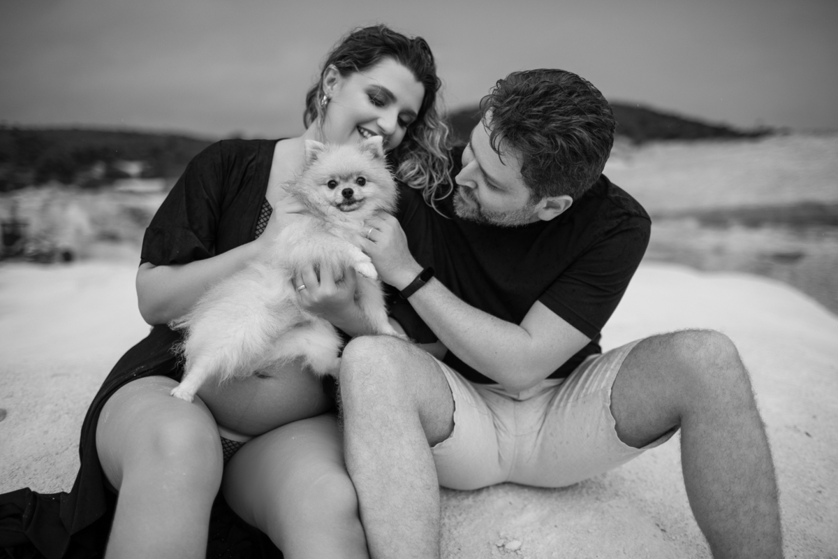 Cena familiar com o casal gestante e o cachorro sentados juntos na areia, olhando para o horizonte. O momento tranquilo reflete o carinho e a união da família. Fotos feitas pelo fotógrafo de gestante Rafael Ohana em Brasília.