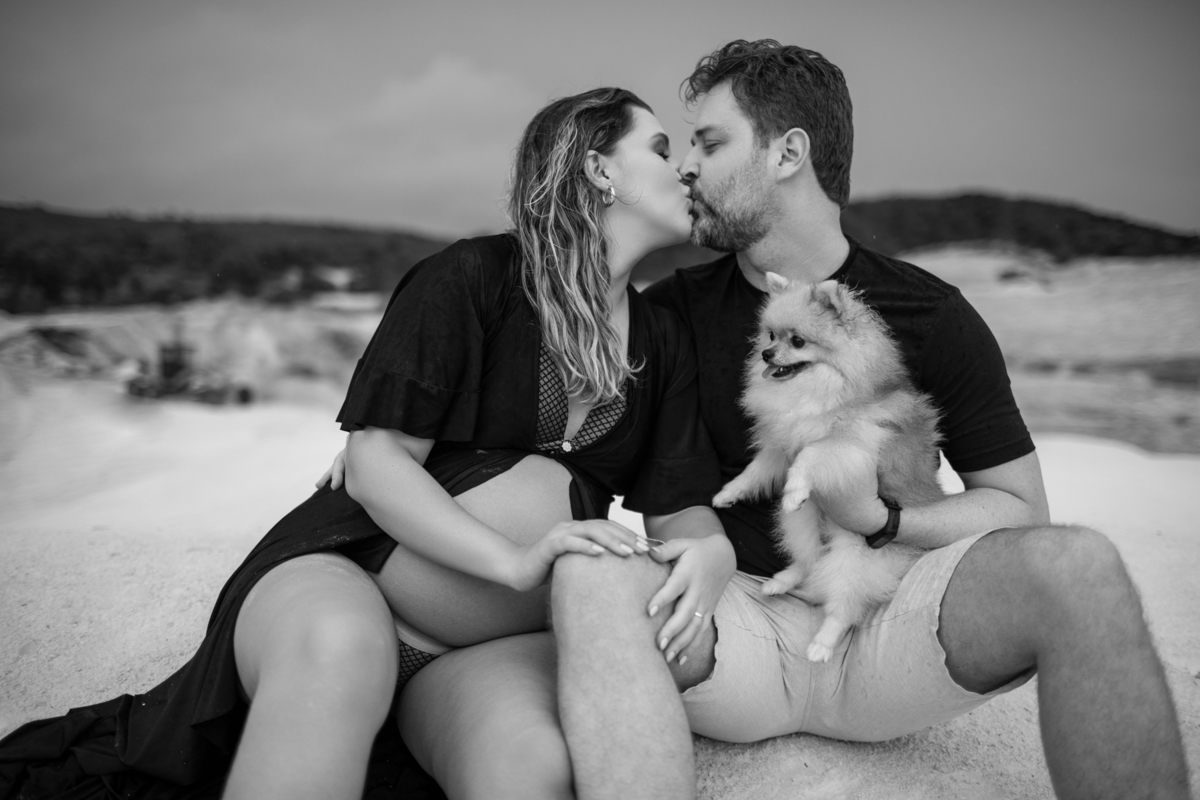 O casal sentado na areia, trocando um beijo carinhoso, enquanto seguram o cachorro no colo. A imagem capta um momento íntimo de afeto, com a tranquilidade do cenário natural ao fundo. Fotos feitas pelo fotógrafo de gestante Rafael Ohana em Brasília.