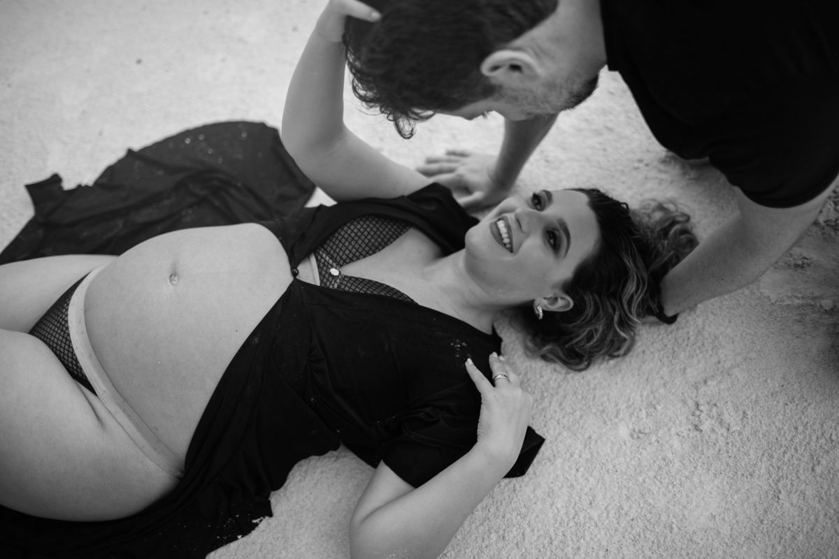 A gestante deitada na areia, sorrindo enquanto o parceiro interage com ela. O ângulo da foto e a expressão dela destacam a felicidade desse momento especial. Fotos feitas pelo fotógrafo de gestante Rafael Ohana em Brasília.