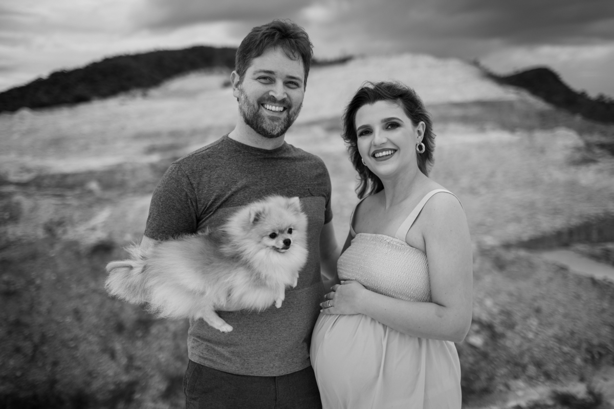 O casal em um momento de carinho, com o homem sorrindo ao lado da gestante. Eles seguram um cachorrinho fofo, criando uma atmosfera familiar e aconchegante. Ao fundo, dunas de areia que completam o cenário. Fotos feitas pelo fotógrafo de gestante Rafael O