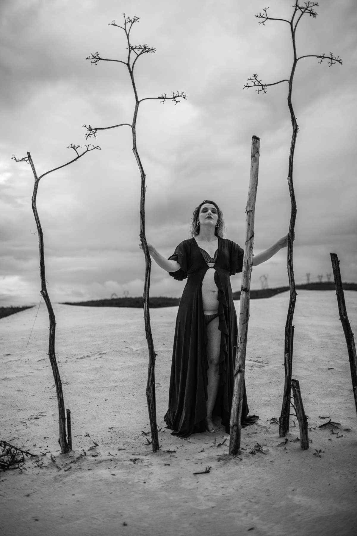 A gestante de pé, entre árvores secas, olhando para o horizonte. A composição dramática das árvores e o céu nublado criam um contraste marcante com a serenidade da figura central. Fotos feitas pelo fotógrafo de gestante Rafael Ohana em Brasília.