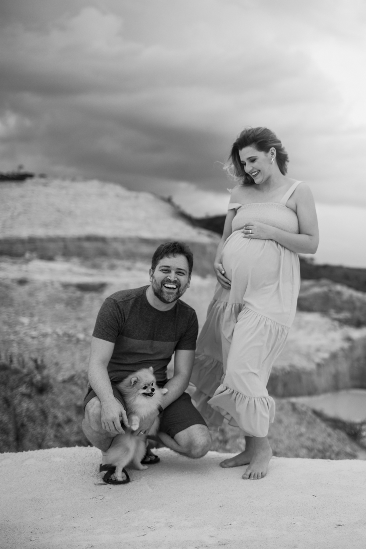 A gestante, em pé, sorrindo, com uma das mãos sobre a barriga e a outra posicionada em seu peito. A expressão de felicidade dela contrasta com a vastidão das dunas ao fundo. Fotos feitas pelo fotógrafo de gestante Rafael Ohana em Brasília