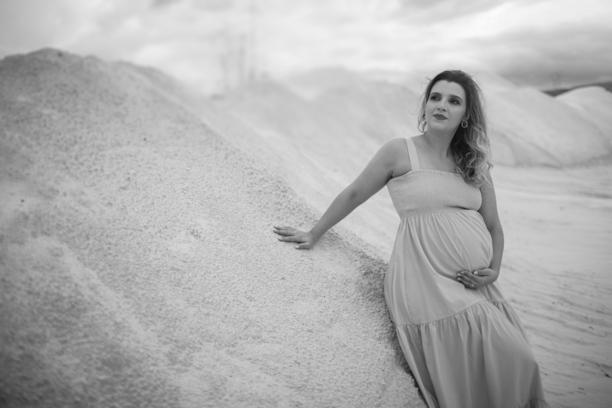 A gestante, em pé, sorrindo, com uma das mãos sobre a barriga e a outra posicionada em seu peito. A expressão de felicidade dela contrasta com a vastidão das dunas ao fundo. Fotos feitas pelo fotógrafo de gestante Rafael Ohana em Brasília