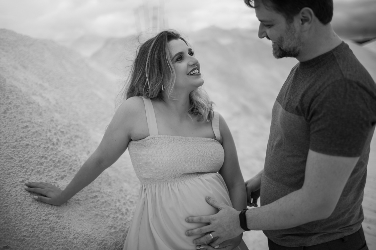 O casal em uma troca de olhares carinhosa. A gestante segura delicadamente a barriga enquanto o parceiro olha para ela, criando uma cena de ternura. As dunas brancas de areia fornecem um pano de fundo dramático. Fotos feitas pelo fotógrafo de gestante Raf