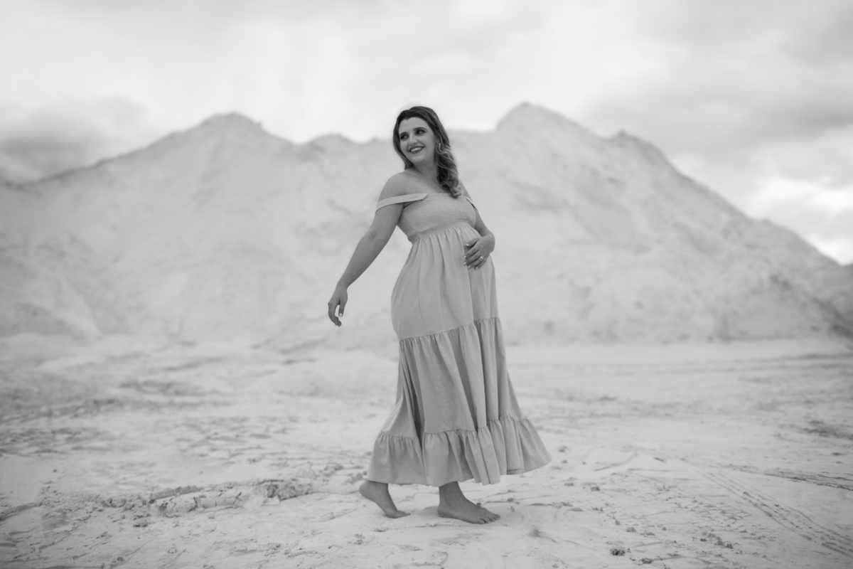 A gestante de pé em meio às dunas brancas, segurando seu vestido leve. O cenário é deslumbrante com as colinas de areia ao fundo, destacando a figura dela no ambiente natural. Fotos feitas pelo fotógrafo de gestante Rafael Ohana em Brasília.