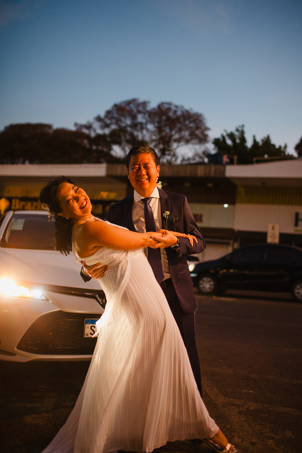 Noivos japoneses em frente a um carro com faróis acesos. Foto feita pelo fotógrafo de casamento Rafael Ohana em Brasília