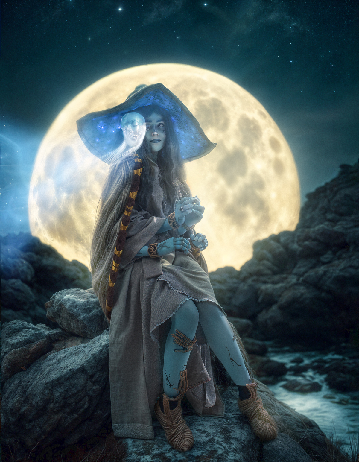 Cosplay de Ranni (Elden Ring). Mulher azulada sob a lua cheia gigante senta-se em rochas, com chapéu estrelado e figura fantasmagórica ao lado. Atmosfera mágica e serena. Foto por Rafael Ohana em Brasília.