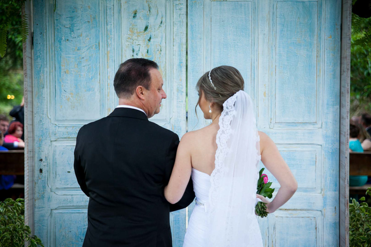 Casamento ao ar livre e de dia dos noivos Anny e Rodolfo fotografado pelo melhor fotógrafo de Campo Largo e região de Curitiba, Michel Druziki. Noiva e pai da noiva em frente à porta da entrada da cerimonia