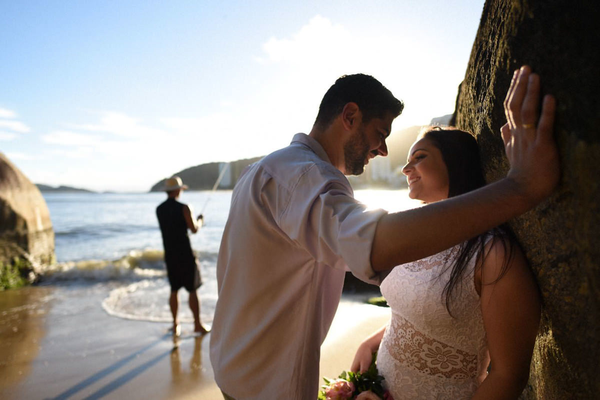 Ensaio pre-wedding dos noivos Vanessa e Adriano. Fotografados por um dos melhores fotógrafos de casamento do Brasil, Michel Druziki. O ensaio foi realizado na praia. Noivos encostados na pedra na areia da praia, pescador ao fundo