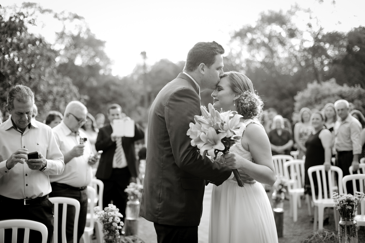 Casamento de dia ao ar livre dos noivos Cheila e Lúcio, no Negrão Eventos, fotografado pelo melhor fotógrafo de casamentos de campo largo e Curitiba, Michel Druziki. Noivas sendo recebida com beijo na testa pelo noivo