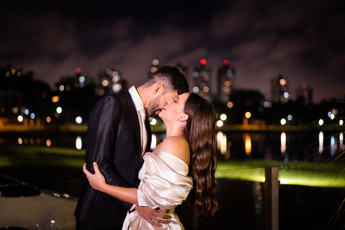 Curitiba Rebeca e Vinicius no Magiiore Barigui fotografado pelo fotógrafo de casamento Michel Druziki. Noivos se beijando em frente a um lago com as luzes da cidade desfocadas