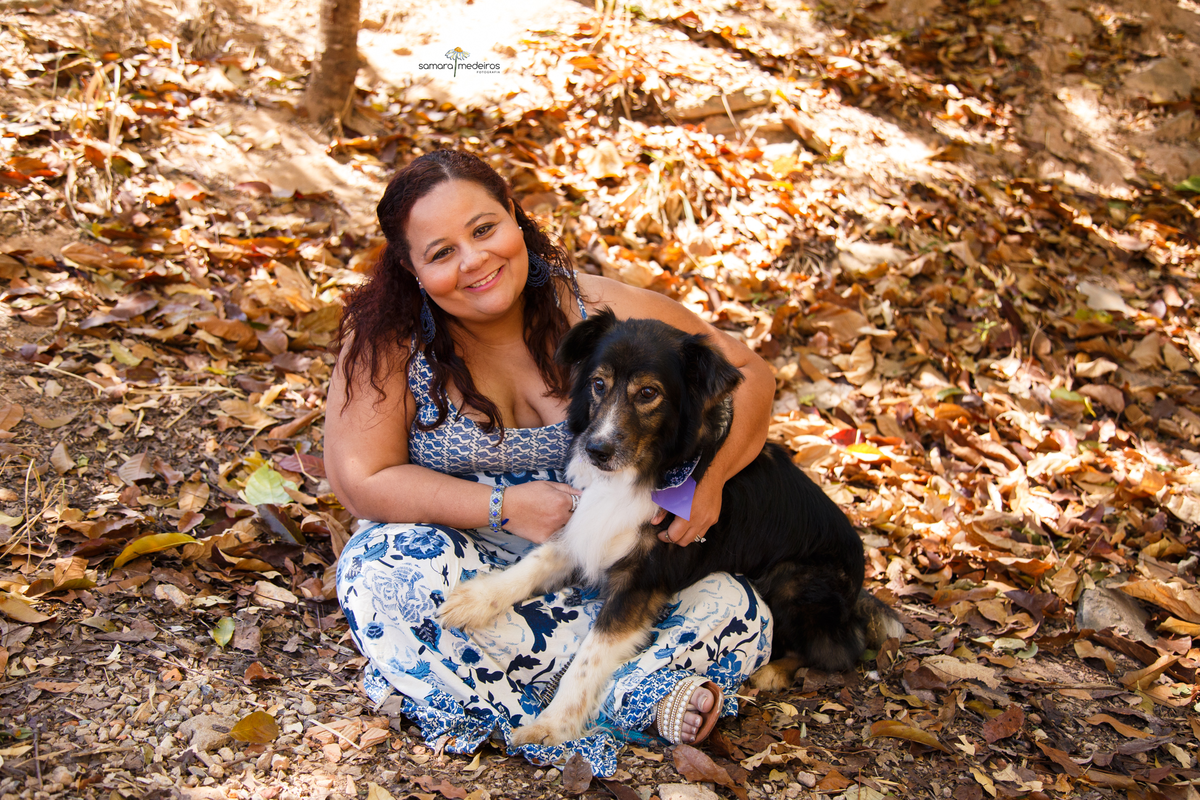 Mulher sentada no chão coberto de folhas com seu cachorro border collie no colo, sorrindo para a câmera.